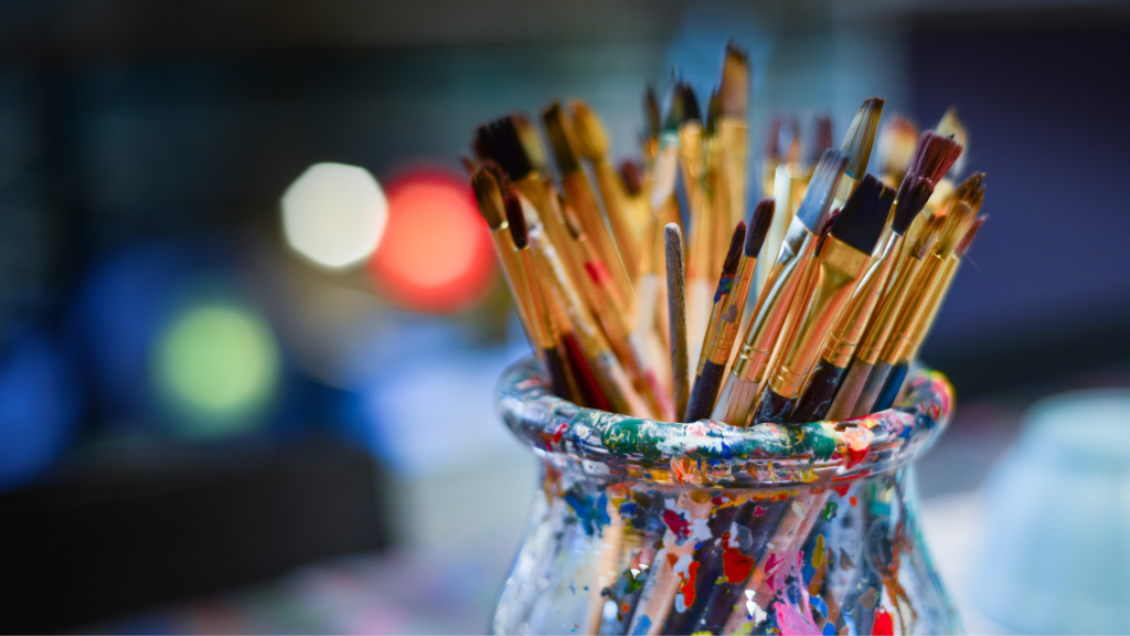 Glass jar covered in specs of paint and filled with paint brushes